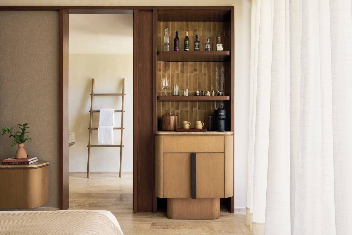 A hotel room with a wooden wardrobe, a small table with a plant, a ladder, and a shelf with bottles and glasses.