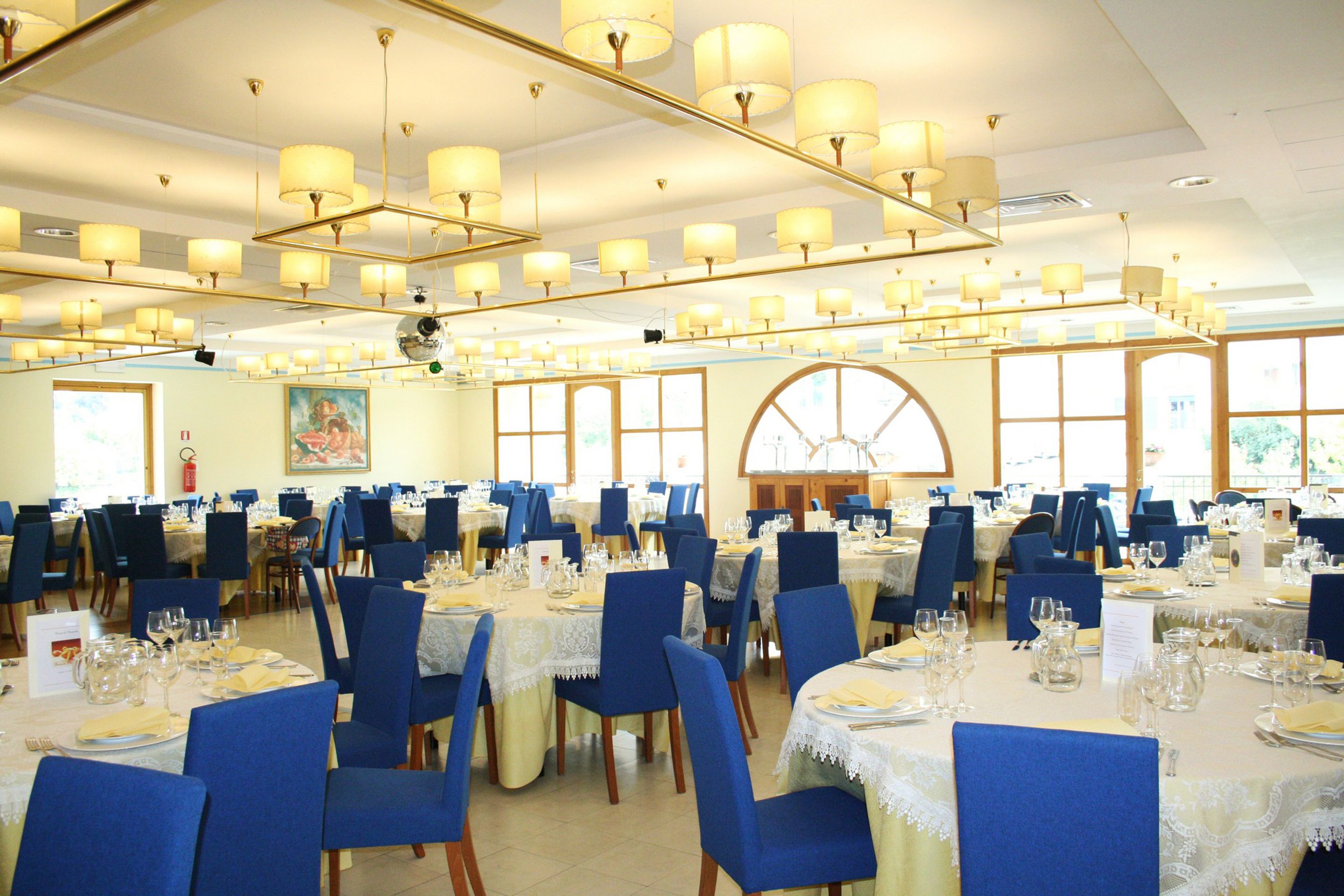 Une grande salle à manger avec de nombreuses tables dressées pour les repas, des chaises bleues et des lustres.