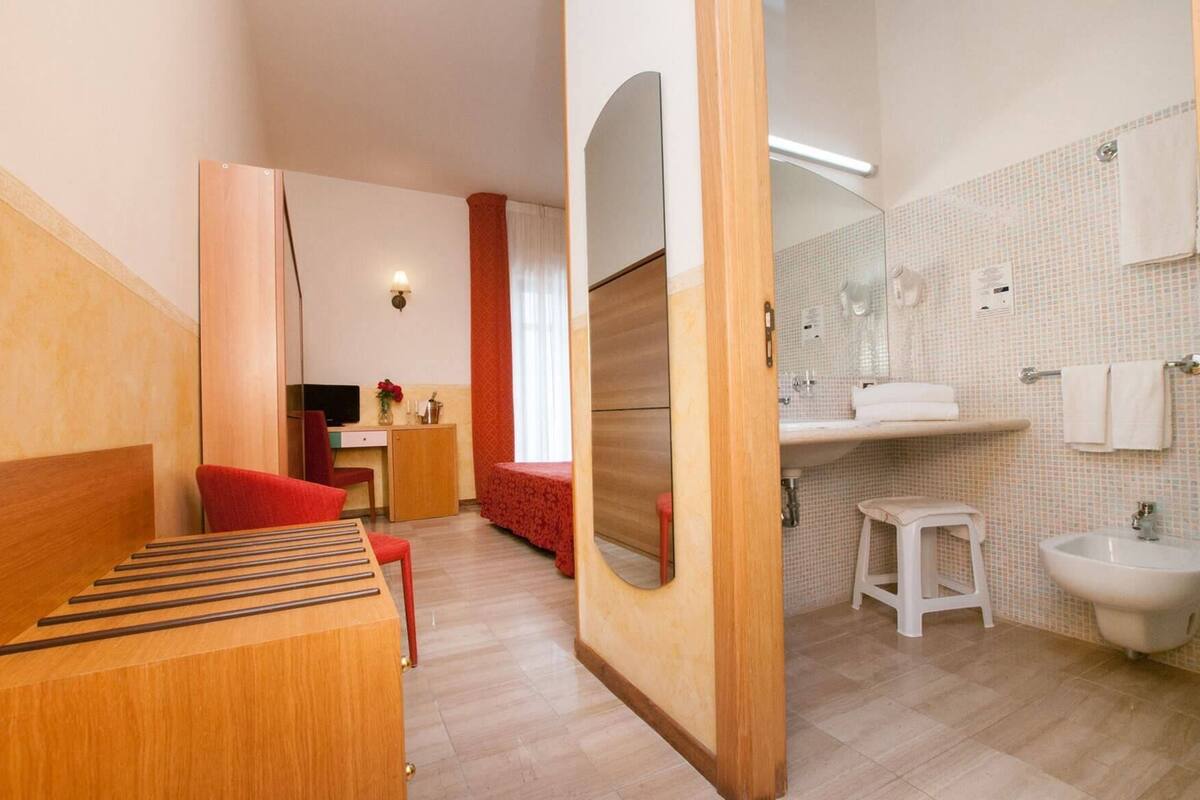 A modern hotel room with a wooden bed, a desk, a red chair, a small table, and a bathroom with a sink and mirror.