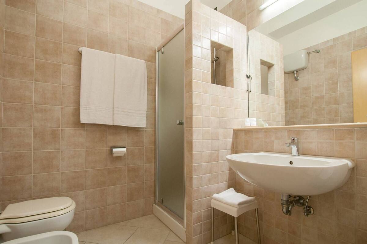 A bathroom with beige tiles, a toilet, a shower, a sink, and a small stool.