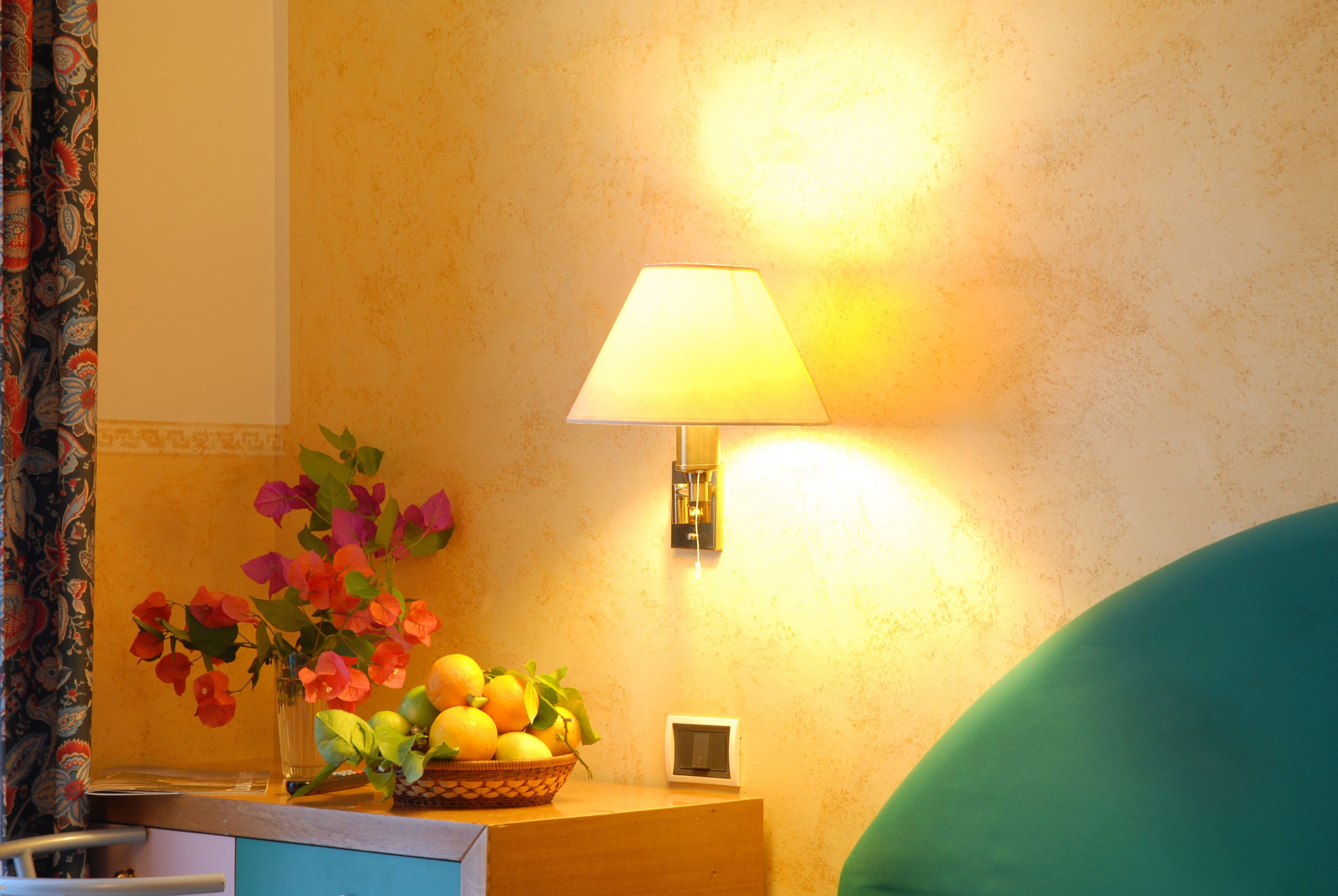 A room with a bedside table holding a vase with flowers and a fruit bowl. There is a lamp on the wall and a chair in the corner.