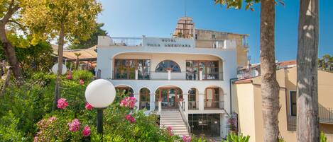 A two-story hotel with a balcony, surrounded by lush greenery and flowering plants.