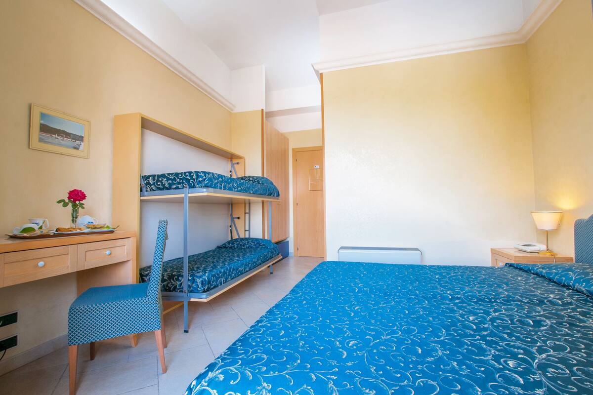 A room with bunk beds, a desk with a chair, a bedside table with a lamp, and a wall-mounted shelf.
