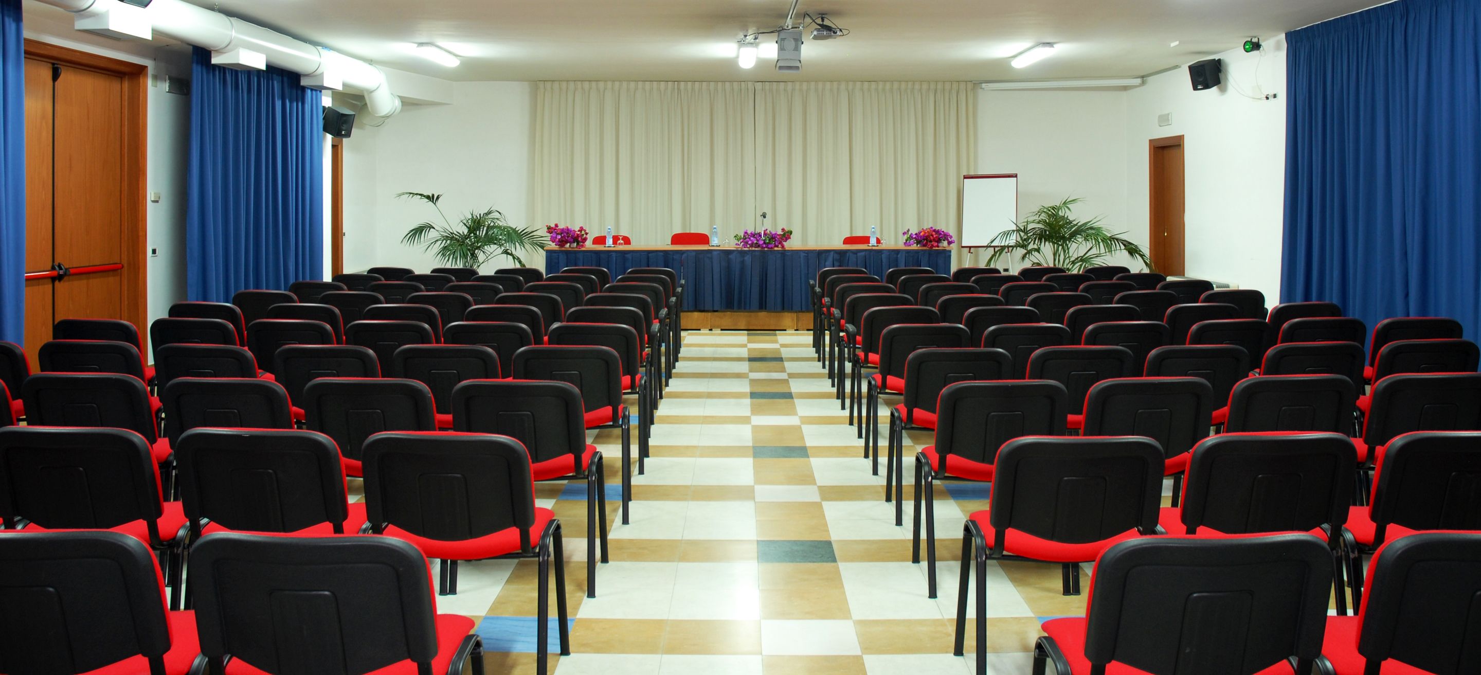 Une salle de conférence avec des rangées de chaises noires dotées de coussins rouges, une scène avec un rideau bleu et un écran de projection.