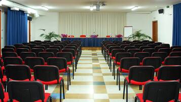 Une salle de conférence avec des rangées de chaises noires dotées de coussins rouges, une scène avec un rideau bleu et un écran de projection.