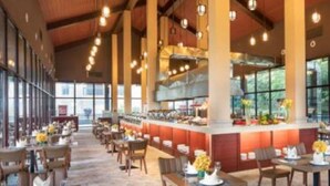 Um restaurante espaçoso com pé direito alto, vigas de madeira e luminárias pendentes. As mesas estão dispostas com toalhas brancas e arranjos de mesa.