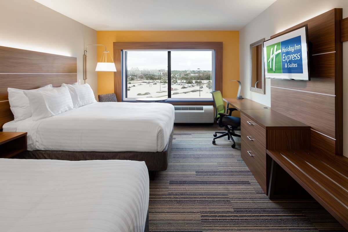 Une chambre d’hôtel avec deux lits, un bureau, une chaise, une grande fenêtre donnant sur un beau paysage et une enseigne indiquant « Holiday Inn Express & Suites ».
