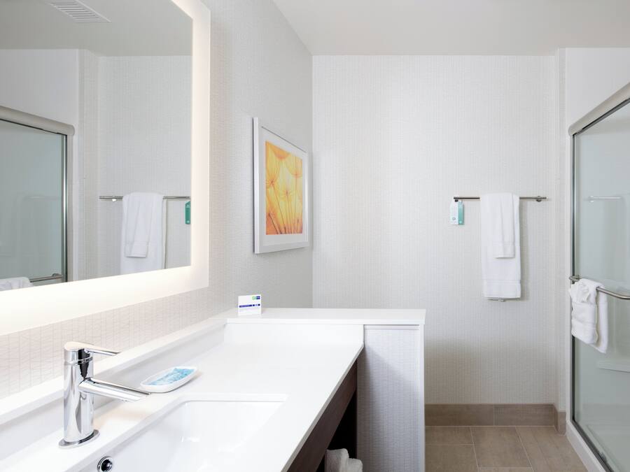 Une salle de bain moderne avec un grand miroir, un lavabo blanc, une douche en verre et une œuvre d’art encadrée au mur.