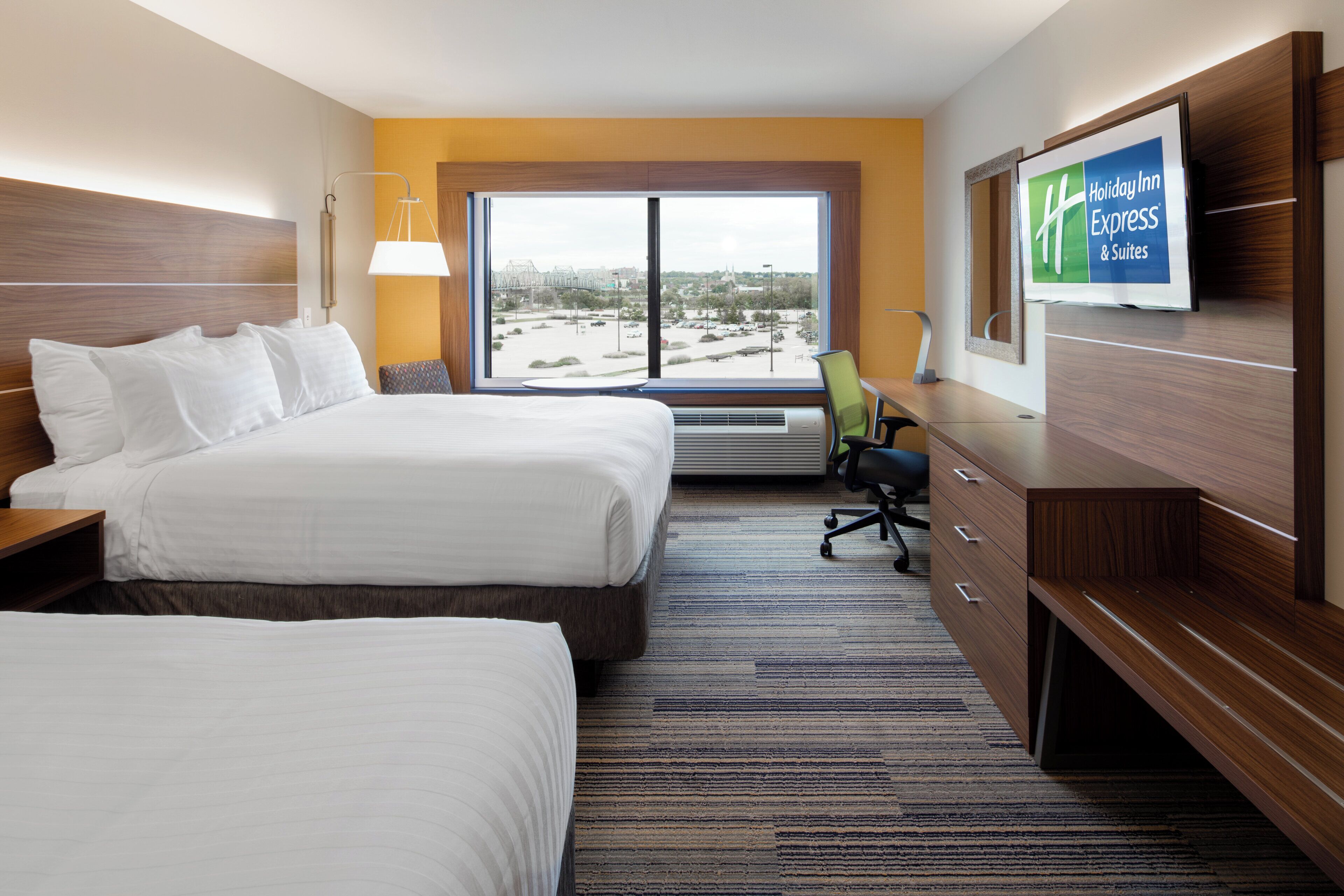 Une chambre d’hôtel avec deux lits, un bureau, une chaise, une grande fenêtre donnant sur un beau paysage et un téléviseur fixé au mur, sur lequel est affiché le logo de Holiday Inn Express.