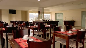 A hotel dining area with multiple tables and chairs, a flat-screen TV mounted on the wall, and large windows offering a view of the city. - Hotel Sollievo (Irati)