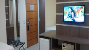 A hotel room with a television mounted on the wall, a small refrigerator, a bed, a desk, and a door with a sign. - Hotel Sollievo (Irati)
