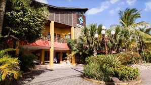 Un hotel de estructura de dos pisos, con balcón y un jardín ajardinado con palmeras y arbustos.
