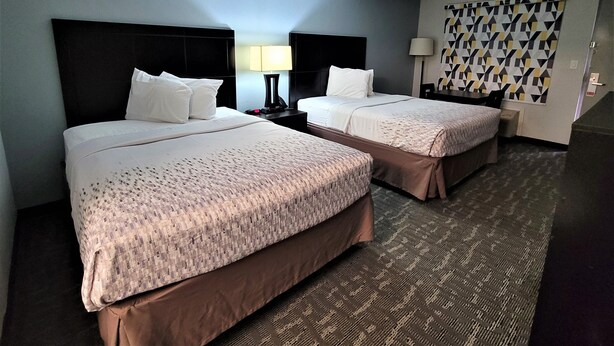 A hotel room with two beds, a nightstand with a lamp, a patterned wall, and a carpeted floor. - Inn of Rockwall (Rockwall)