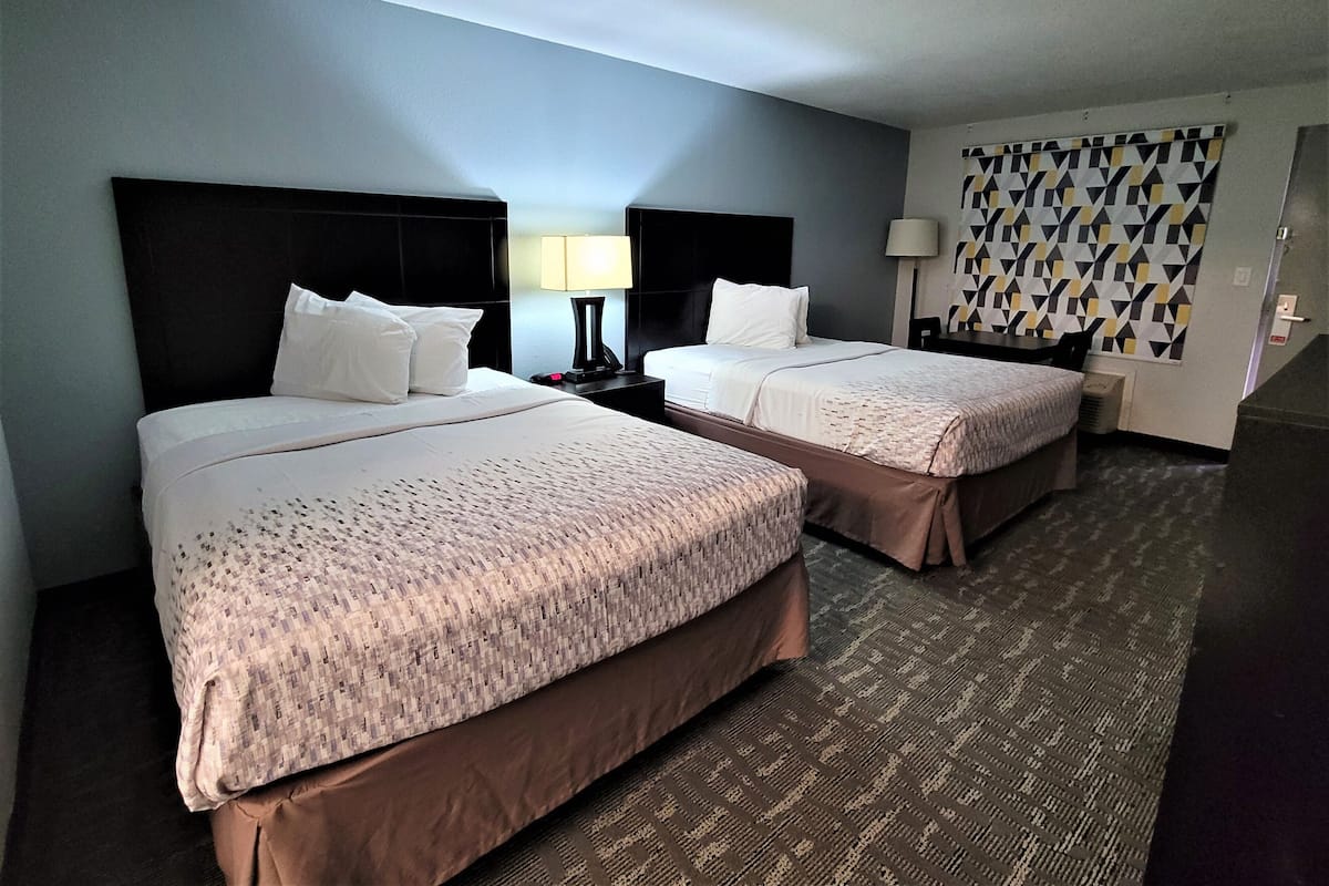 A hotel room with two beds, a nightstand with a lamp, a patterned wall, and a carpeted floor.