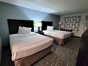 A hotel room with two beds, a nightstand with a lamp, a patterned wall, and a carpeted floor. - Inn of Rockwall (Rockwall)