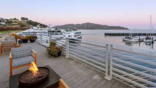 A waterfront deck with outdoor seating, a fire feature, and boats docked in the background. - Waters Edge Hotel (Tiburon)