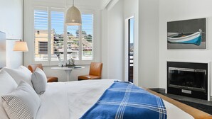 A hotel room with a bed, a table with chairs, a fireplace, and a large window with blinds. - Waters Edge Hotel (Tiburon)