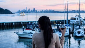 A woman holding a glass of wine, overlooking a marina with boats and a city skyline in the background. - Waters Edge Hotel (Tiburon)