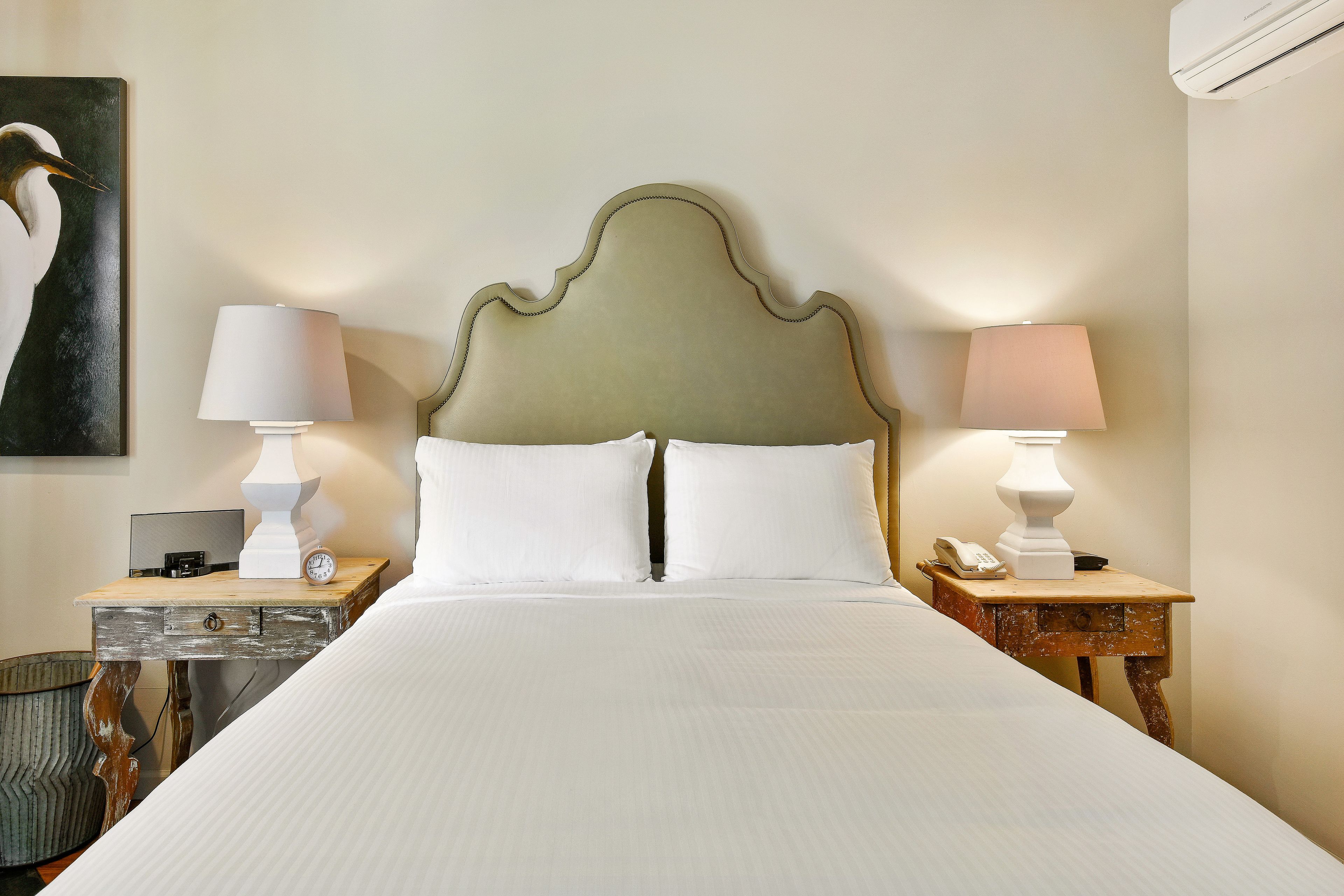 A neatly made bed with white linens, two bedside tables with lamps, a wall-mounted telephone, and a painting of a bird.
