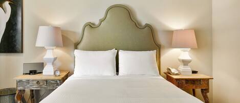 A neatly made bed with white linens, two bedside tables with lamps, a wall-mounted telephone, and a painting of a bird.