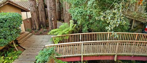 Un pont en bois enjambe un sentier de jardin menant à une maison dont le toit est recouvert de tuiles.