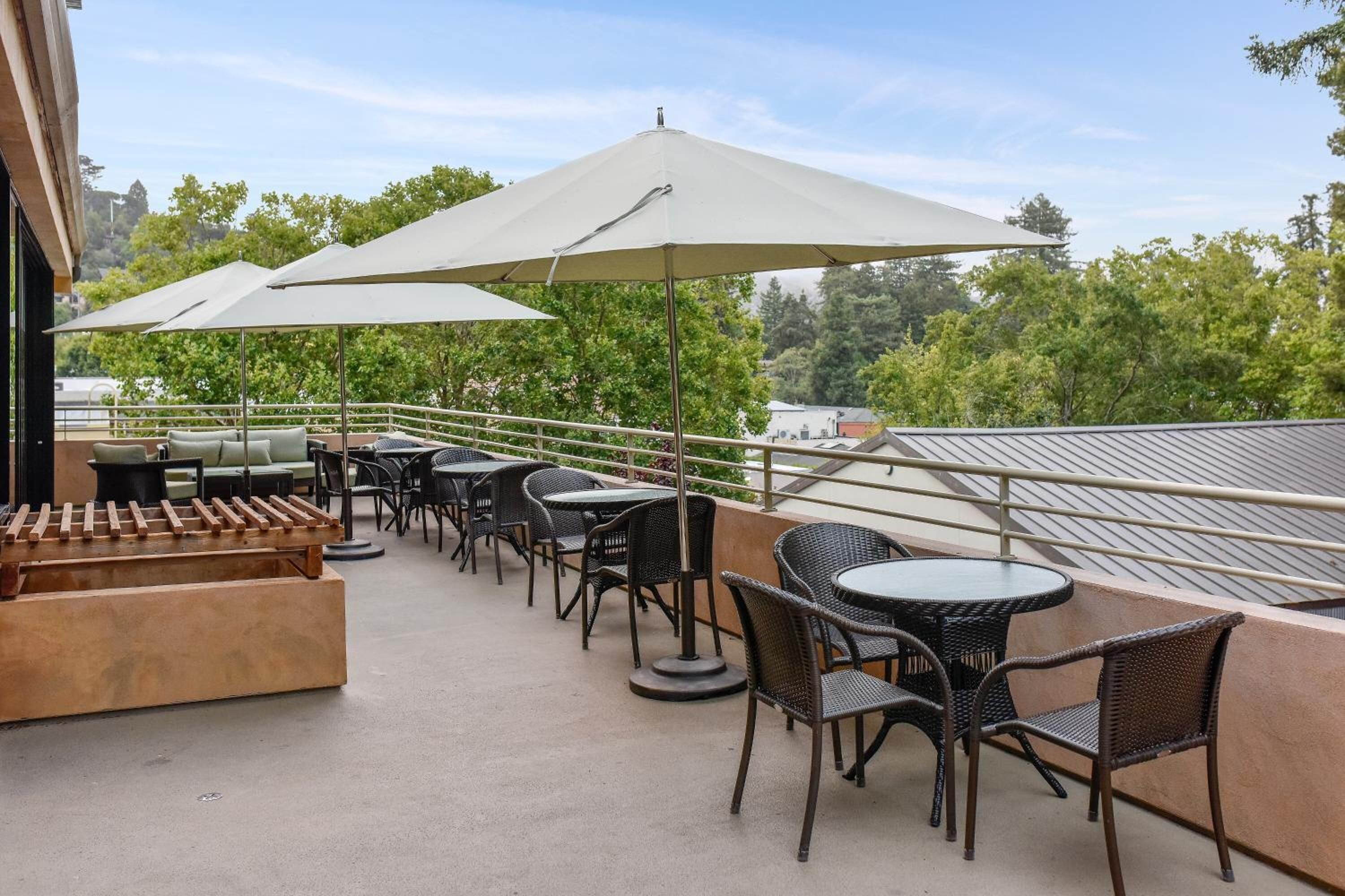 Une terrasse extérieure aménagée avec des meubles en osier, des parasols et offrant une vue sur les arbres et les bâtiments.