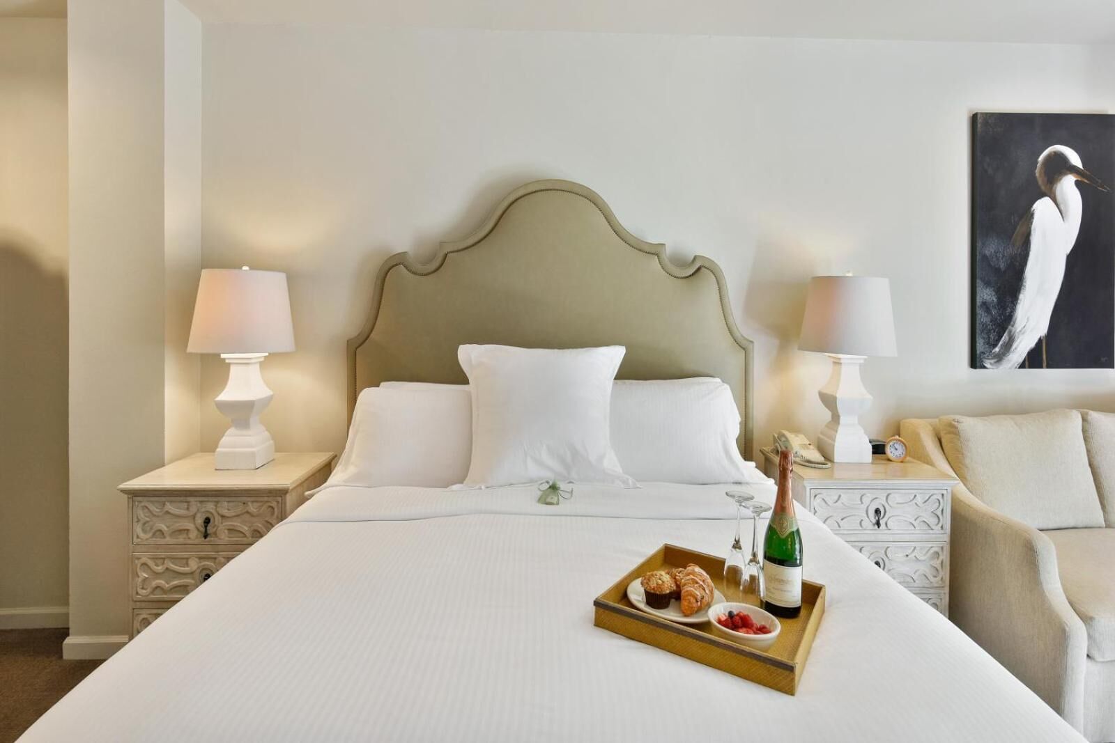 A hotel room with a large bed, two bedside tables with lamps, a nightstand, a tray with food and drink, and a painting on the wall.