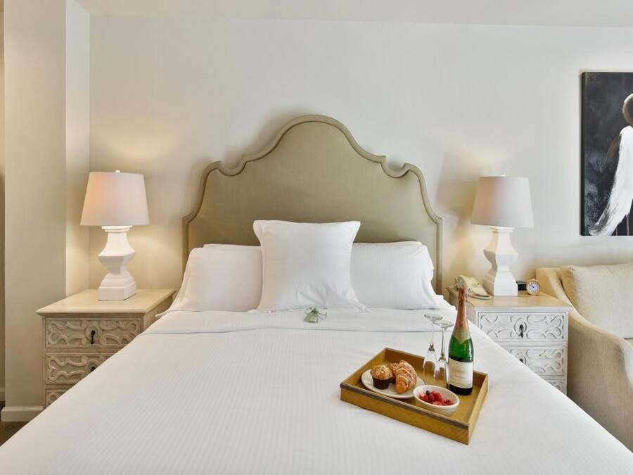 A hotel room with a large bed, two bedside tables with lamps, a nightstand, a tray with food and drink, and a painting on the wall.