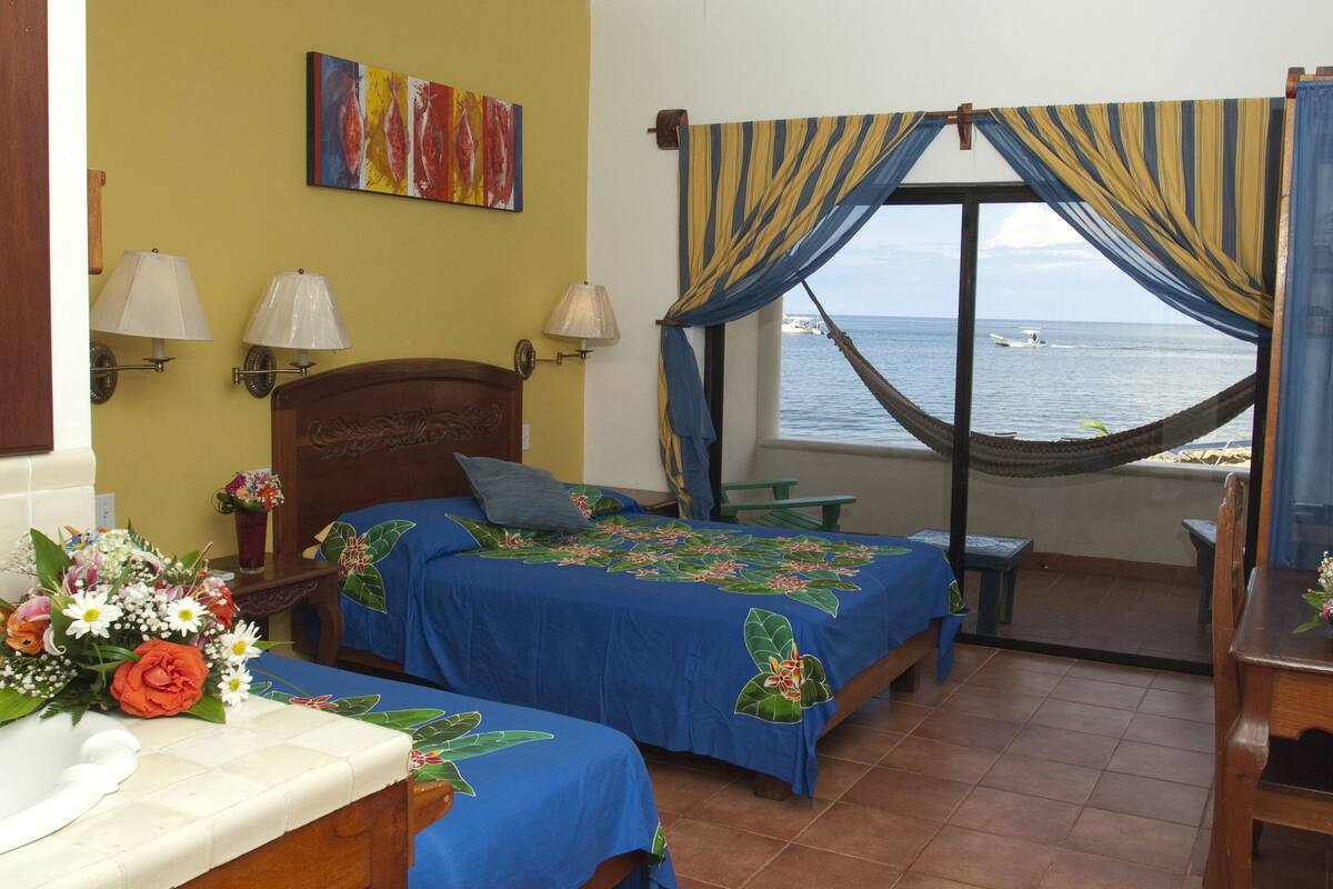 A room with two beds, a view of the sea, a hammock, and a bathroom with a sink and decorative tiles.