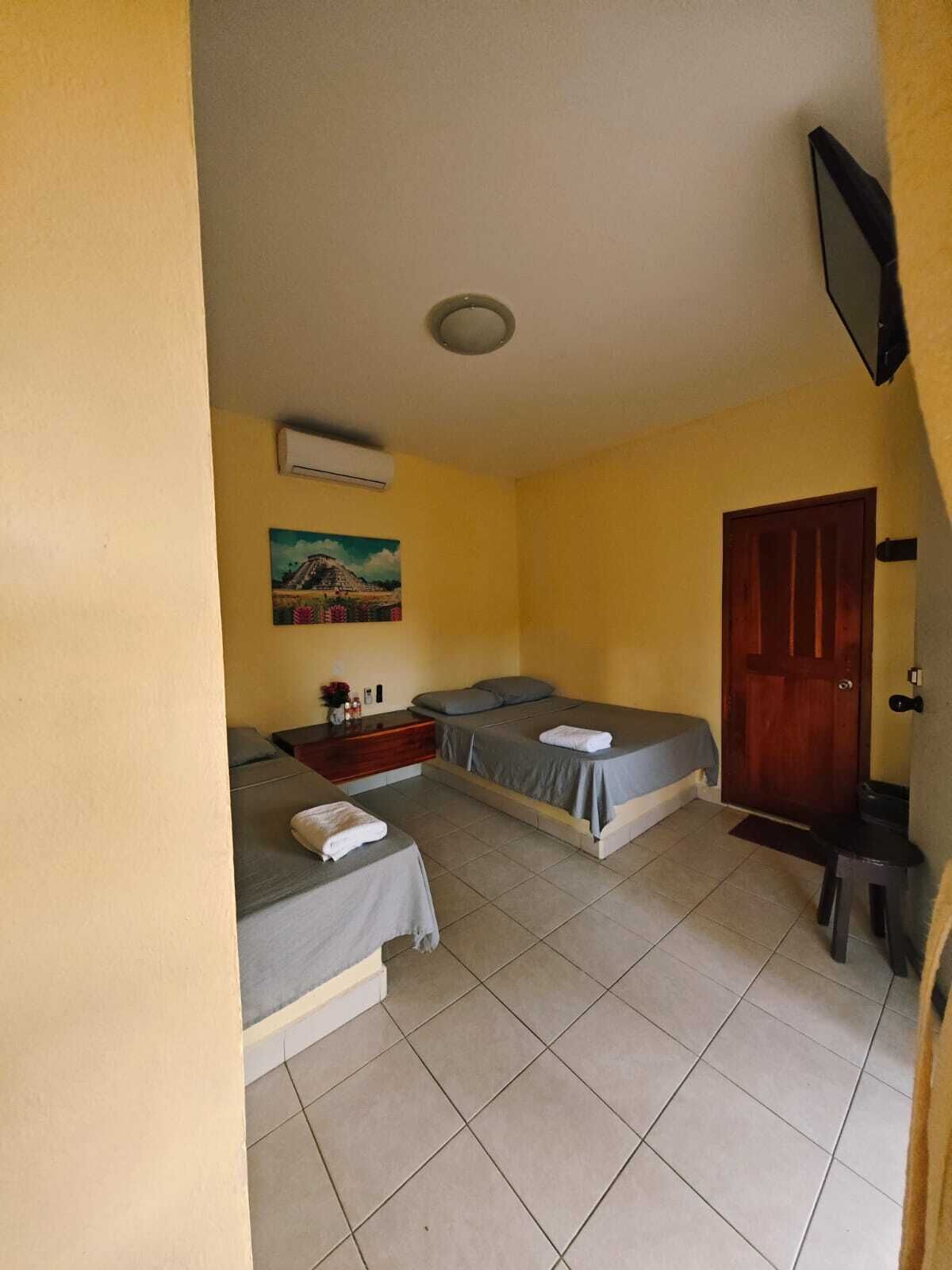 A room with two beds, a small table with a vase, a painting on the wall, and a door.