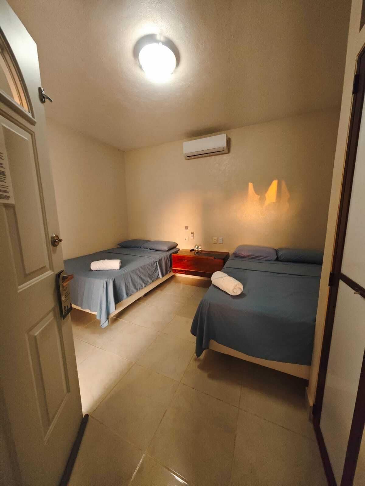A small, well-lit room with two single beds, a bedside table, a wall-mounted air conditioner, and a door with a handle.