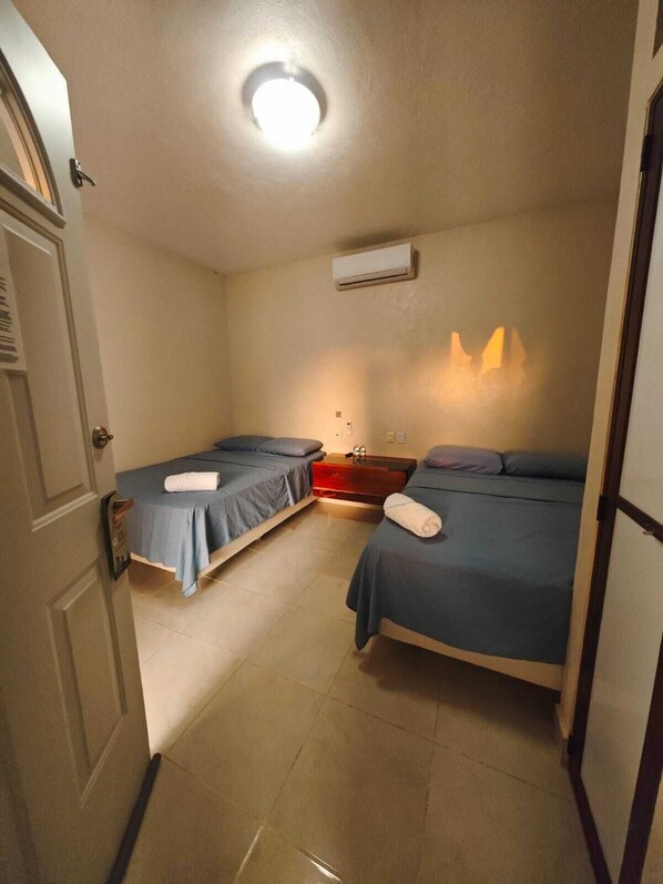 A small, well-lit room with two single beds, a bedside table, a wall-mounted air conditioner, and a door with a handle.