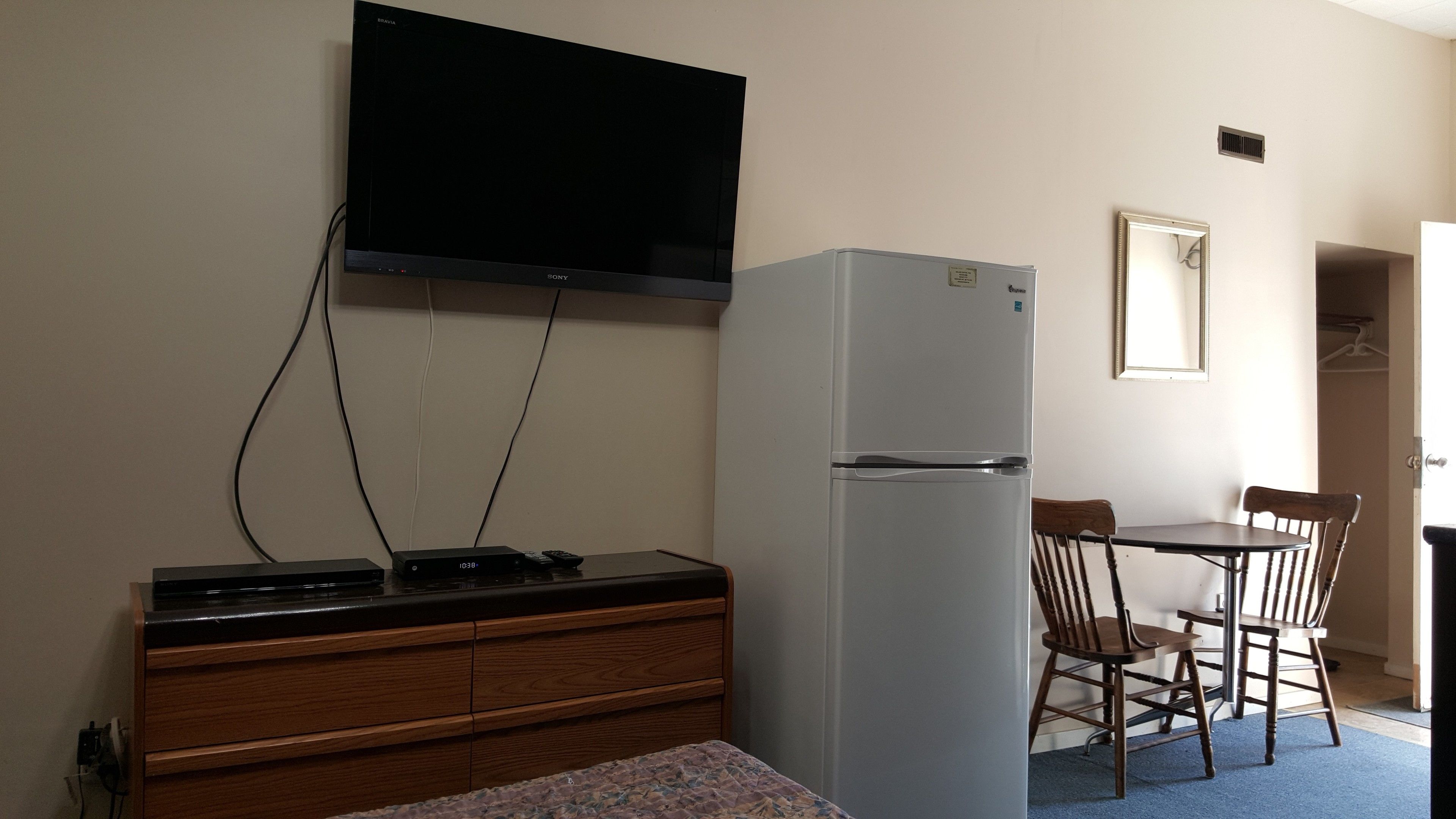 Une pièce compacte avec un téléviseur à écran plat fixé au mur, un réfrigérateur blanc, une commode en bois, une petite table avec deux chaises et une porte donnant sur une autre pièce.