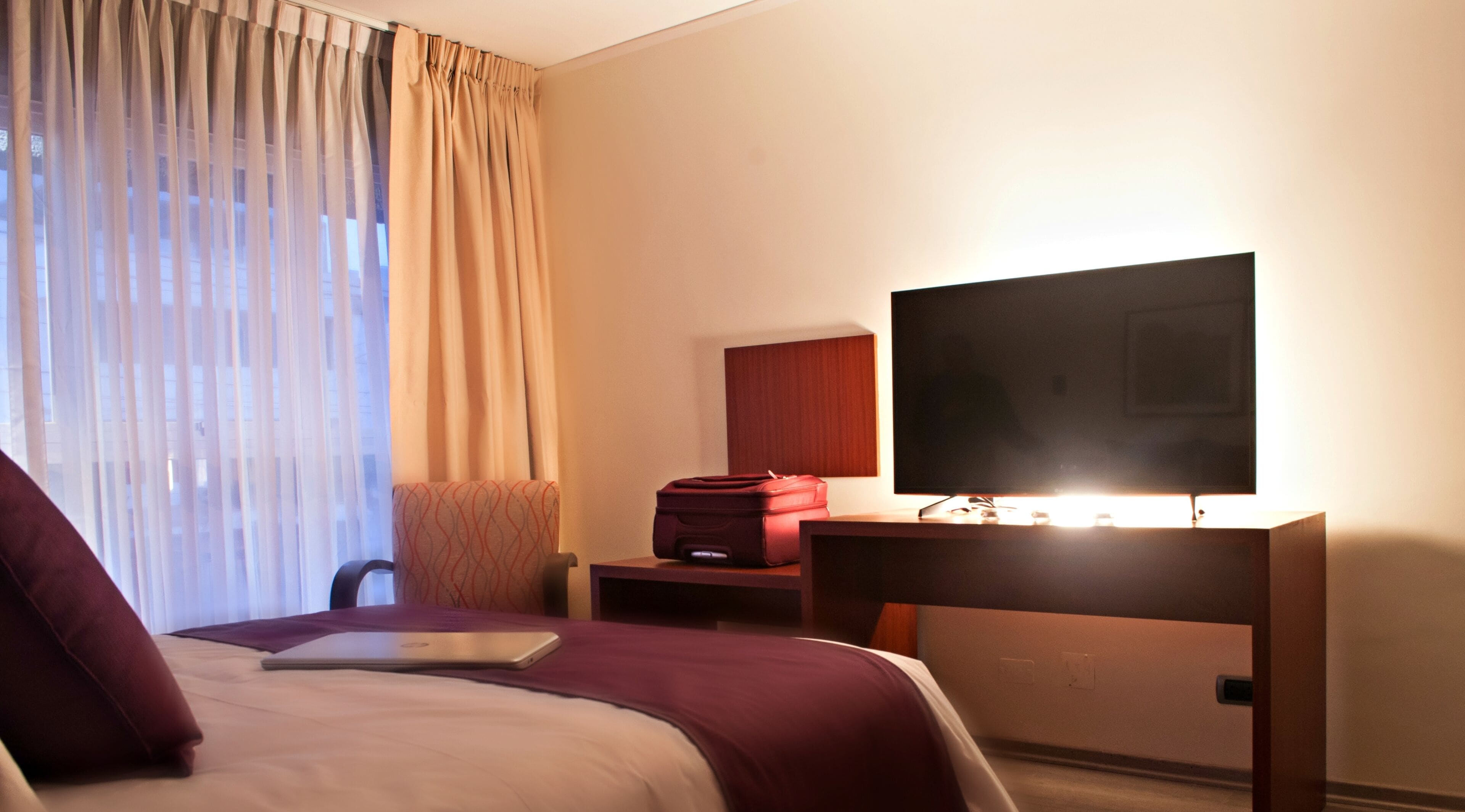 Una habitació d'hotel amb un llit, un escriptori amb televisió, una cadira i una maleta.