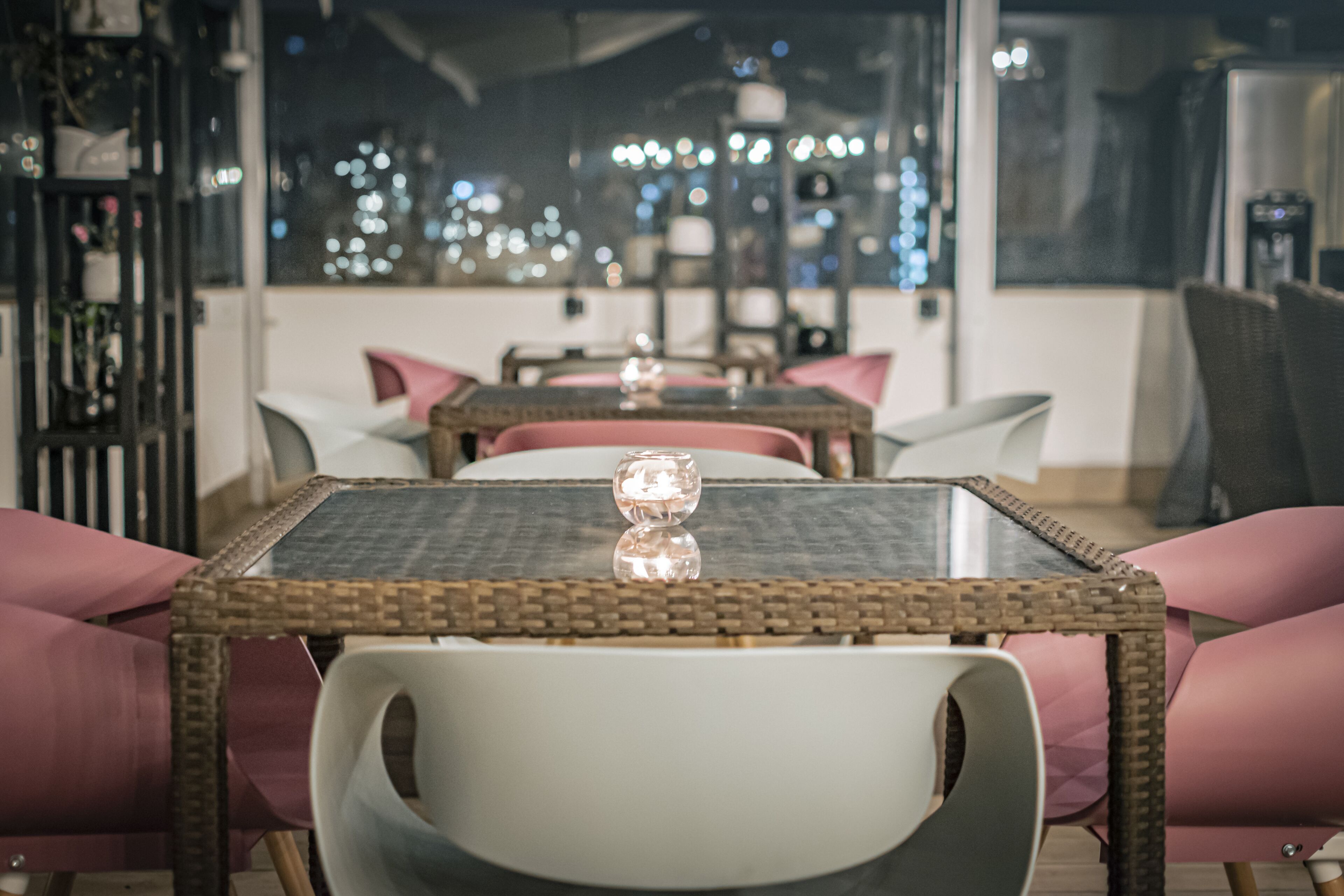 A dining area with wicker tables and cushioned chairs, a decorative glass centerpiece, and a view of city lights through large windows.