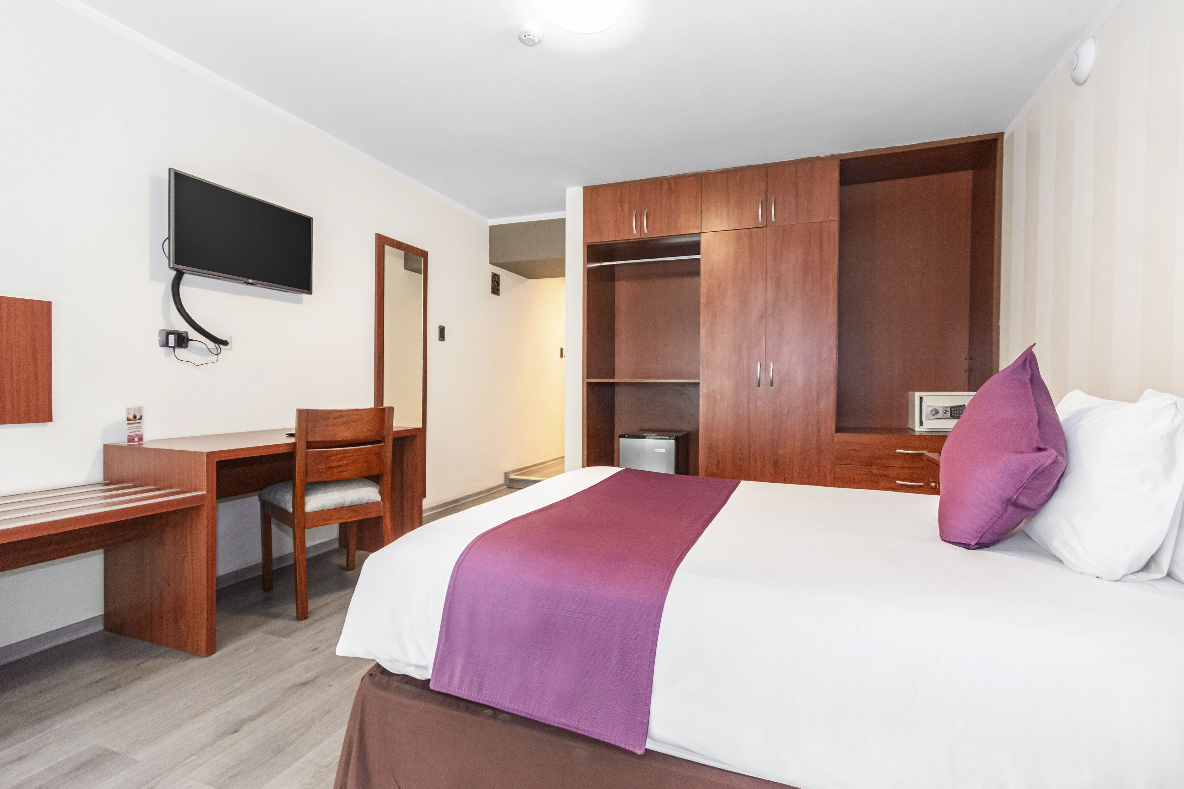 A hotel room with a bed, a desk, a chair, a TV mounted on the wall, and a wardrobe.