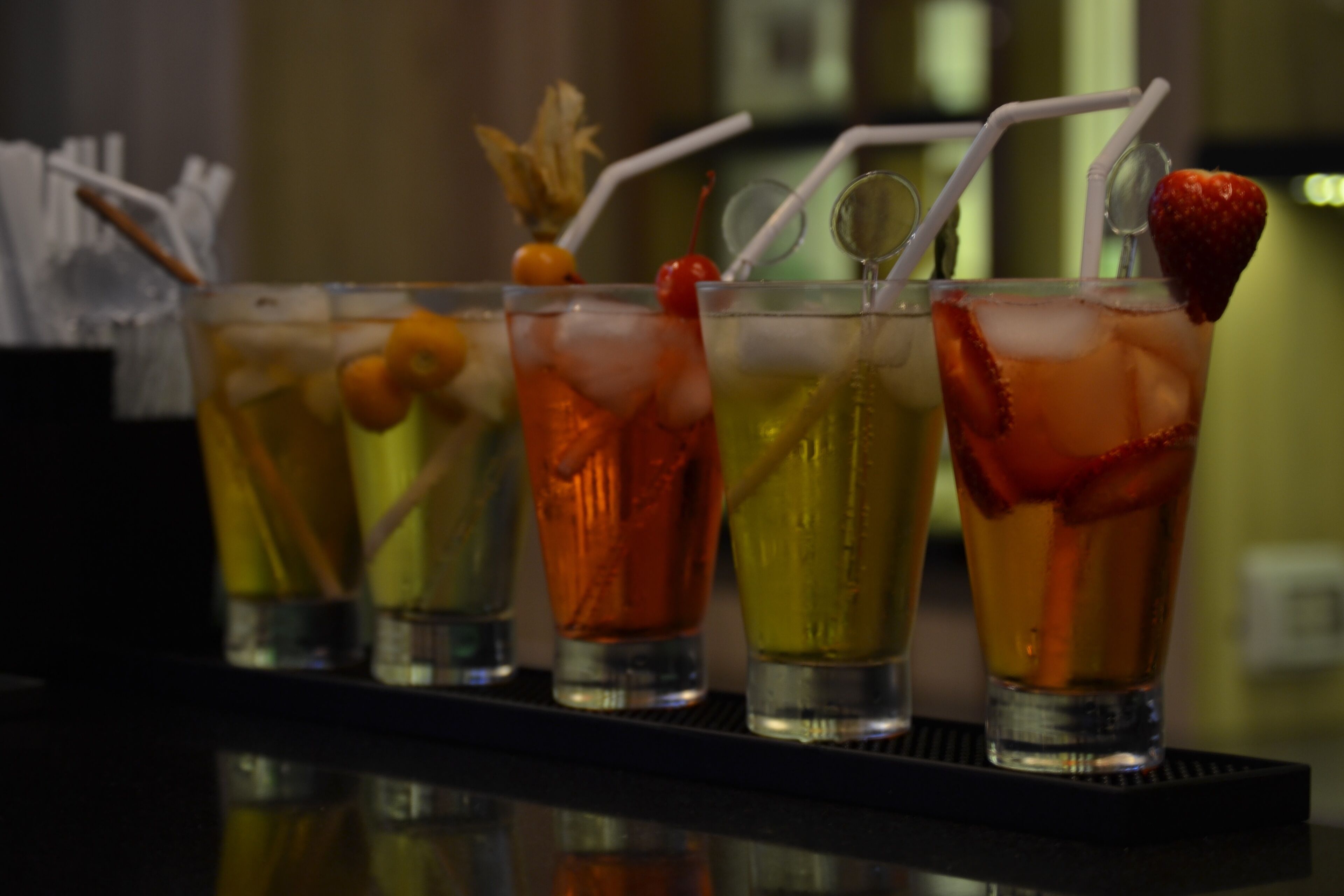 Quatro copos de cocktails com canudos e guarnições sobre um balcão de bar.