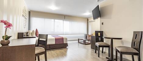 Habitación de hotel con cama, escritorio, dos sillas, televisor montado en la pared y un ventanal grande.