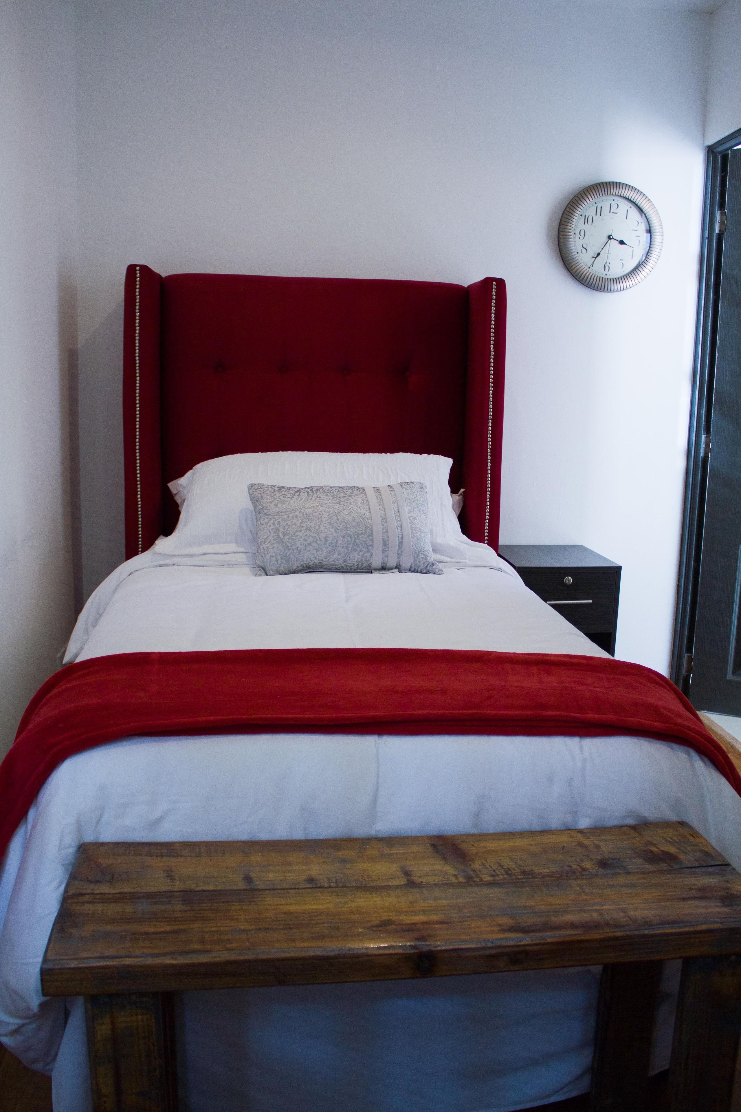 A bed with a red headboard, white bedding, and a grey pillow. There is a wooden bench at the foot of the bed. A clock is mounted on the wall to the right of the bed.