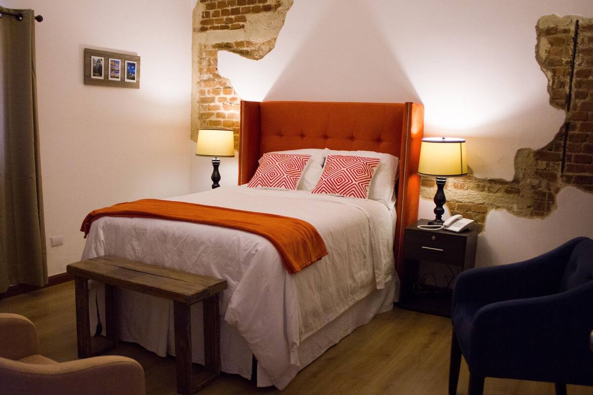 Habitación de hotel con pared de ladrillo, una cama con cabecera naranja, dos lámparas de noche, una mesita de noche de madera, una silla azul y una pequeña mesa.