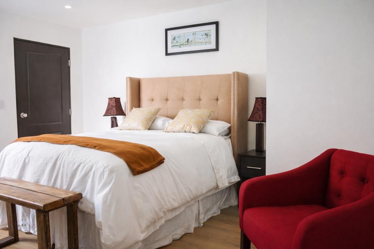 Une chambre à coucher avec un lit, des tables de chevet, une chaise rouge, un banc en bois et une porte.