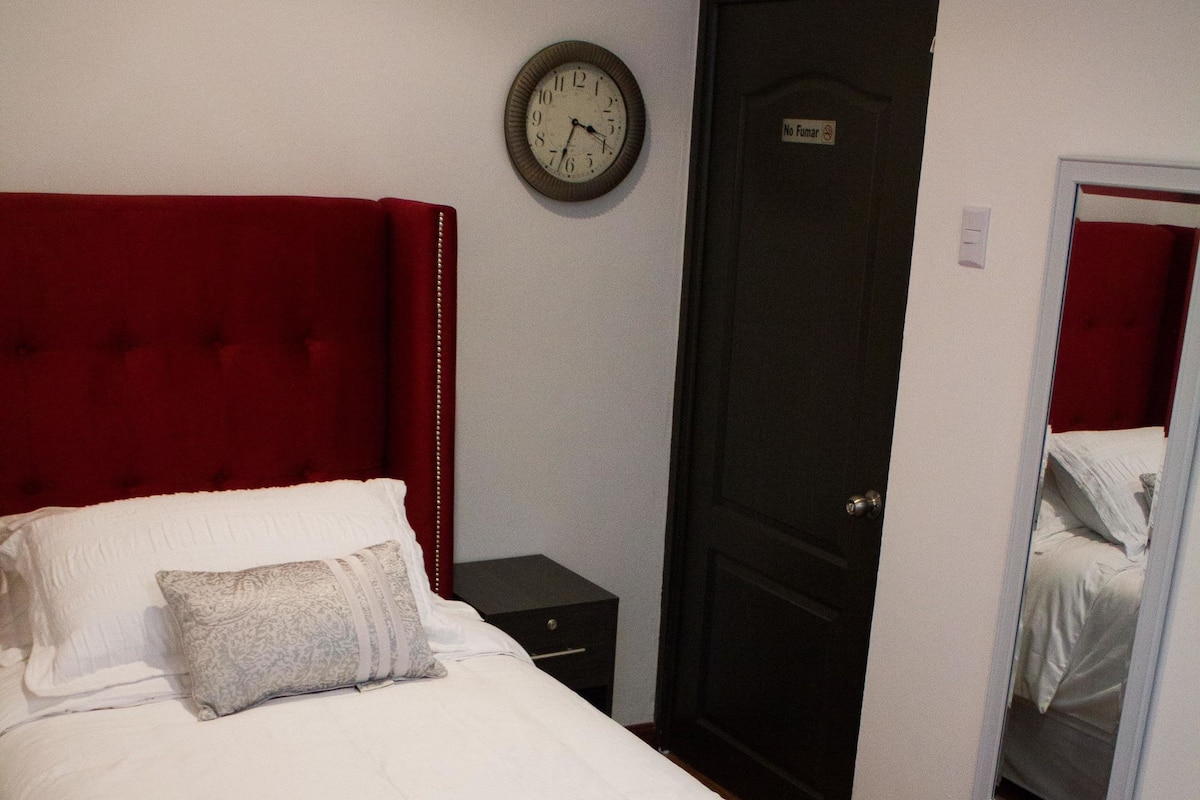Una cama bien hecha con cabecera roja, una mesita de noche, un banco de madera, un reloj y una puerta con un cartel.
