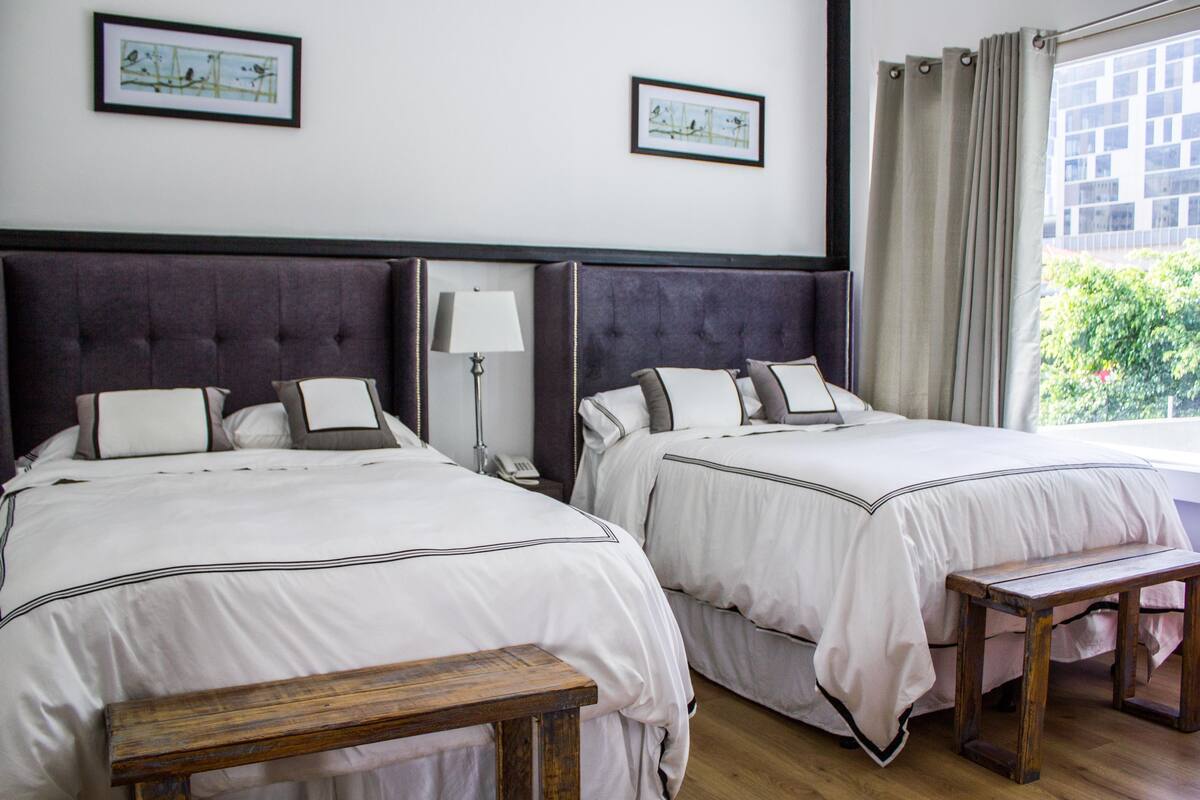 Habitación de hotel con dos camas, una mesita de noche, una lámpara, un banco de madera y una ventana con cortinas.