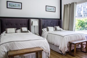 Habitación de hotel con dos camas, una mesita de noche, una lámpara, un banco de madera y una ventana con cortinas.