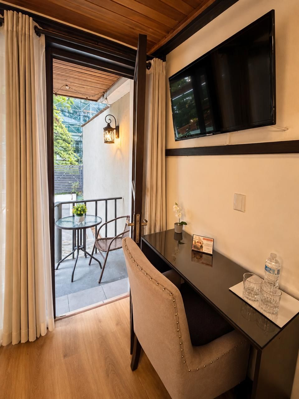 A room with a wooden ceiling, a TV mounted on the wall, a desk with a chair, and a small table with a glass and a plant.