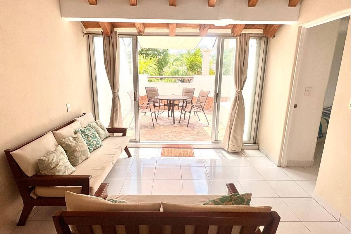 Quarto com sofá, mesa de centro e mesa de jantar com cadeiras. Há uma porta de vidro deslizante que dá para uma área de pátio externo.