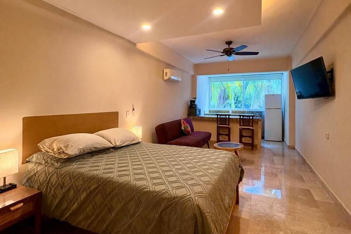 Quarto com cama, escrivaninha, cadeira, televisão e ventoinha no teto.