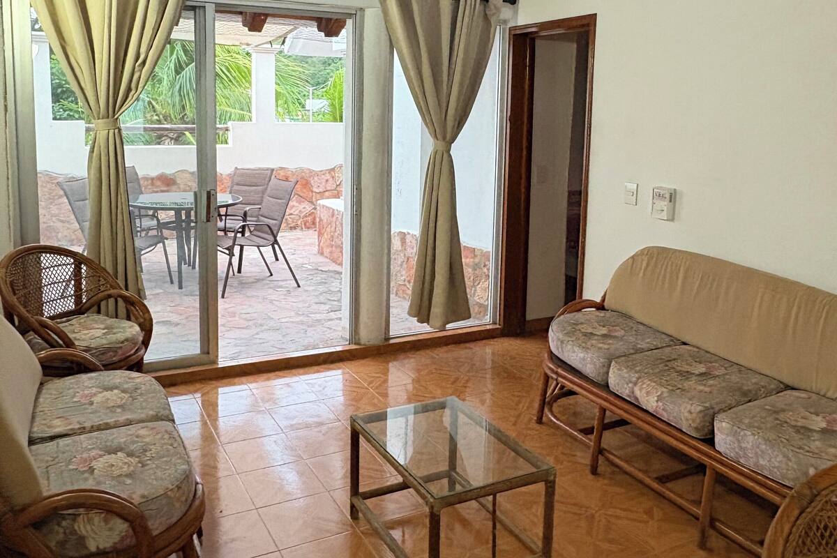 Quarto com ventilador de teto, móveis de vime, mesa de centro de vidro e vista para um pátio externo através de uma porta de vidro.