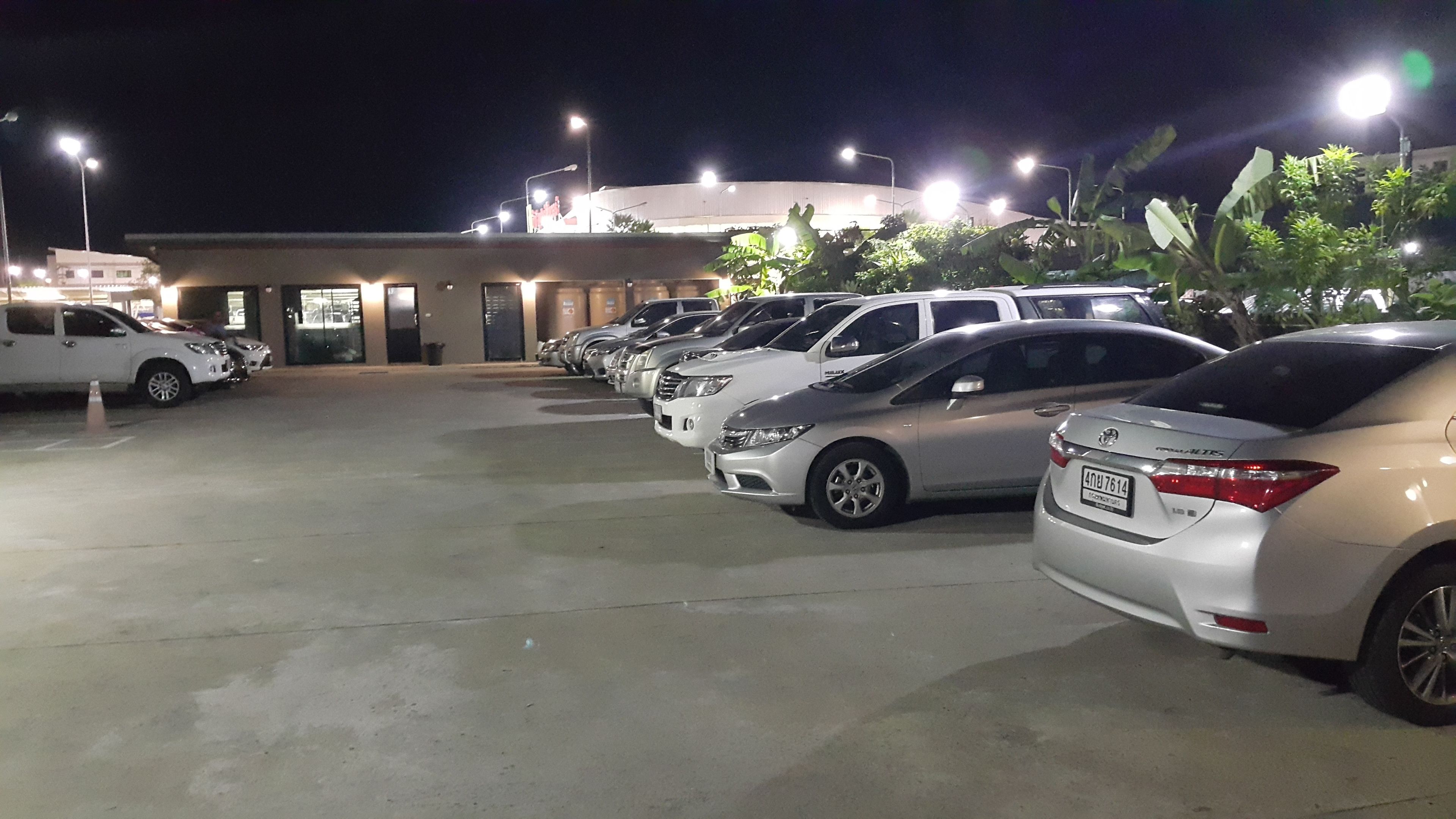 Un estacionamiento con varios coches aparcados en filas, un edificio con ventanas iluminadas y una calle bien iluminada.