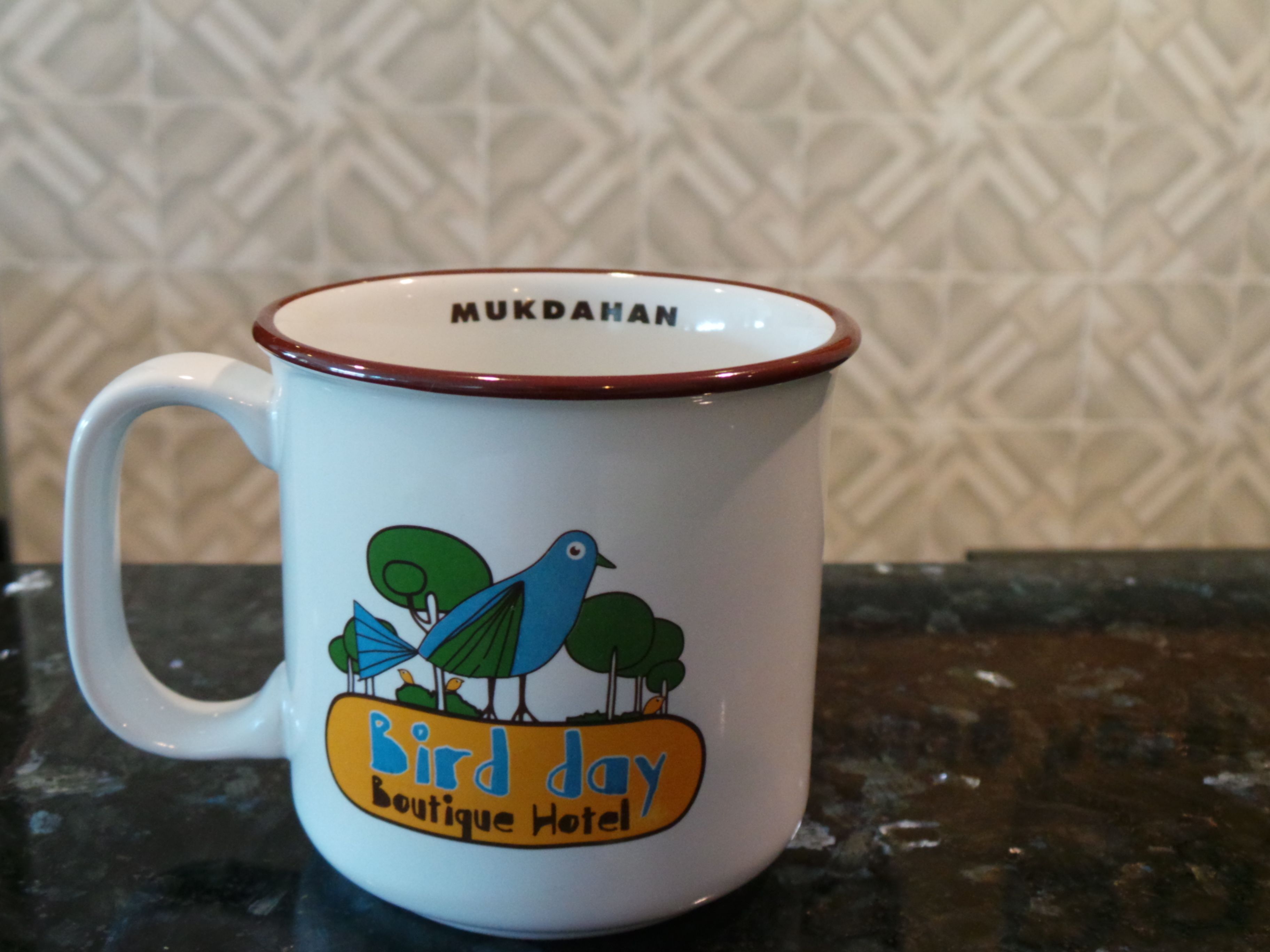 Una taza blanca con borde marrón y un diseño colorido que muestra aves y árboles, con la etiqueta 'Bird day Boutique Hotel'.